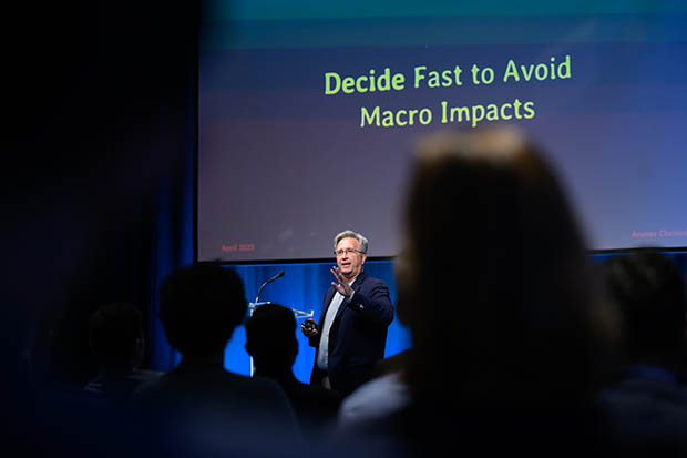 A man speaks to an audience. Behind him, the words "Decide Fast to Avoid Macro Impacts" are displayed on a screen.